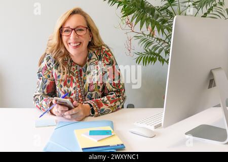 Una allegra donna d'affari bionda con gli occhiali seduti alla sua scrivania, che tiene uno smartphone con un grande schermo del computer nelle vicinanze Foto Stock