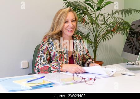 Un'allegra donna d'affari bionda che indossa una giacca colorata si siede alla sua scrivania per rivedere documenti con occhiali da vista nelle vicinanze Foto Stock