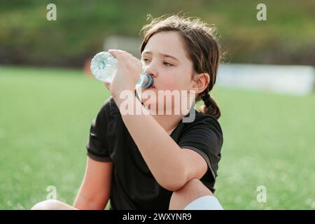 Una ragazza si prende una pausa per bere l'acqua di una bottiglia mentre riposa su un campo da calcio, mostrando una sana idratazione durante lo sport Foto Stock