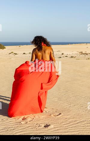 La vista posteriore di una donna irriconoscibile e aggraziata in un abito rosso che scorre cammina sulle dune sabbiose del deserto, guardando lontano dalla telecamera, con il mare sereno in mezzo Foto Stock