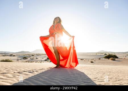 Donna elegante in un abito rosso che scorre posa sulle dune del deserto con gli occhi chiusi, crogiolandosi alla luce del sole dorato. Foto Stock