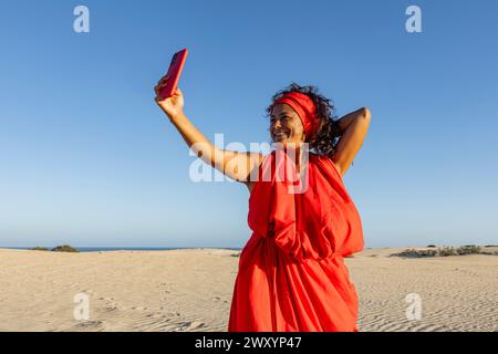 Una donna allegra in un vivace abito rosso si fa un selfie con il suo cellulare, sorridendo ampiamente sulle dune sabbiose del deserto sotto un cielo azzurro. Foto Stock