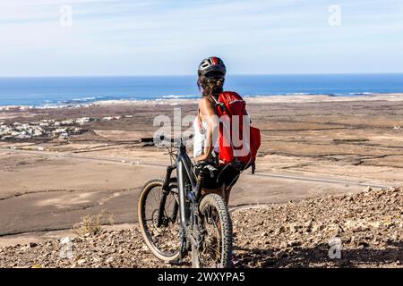 Una donna di mountain bike si erge accanto alla sua bici su una collina, guardando verso il mare e un paesaggio lontano sotto un cielo limpido Foto Stock