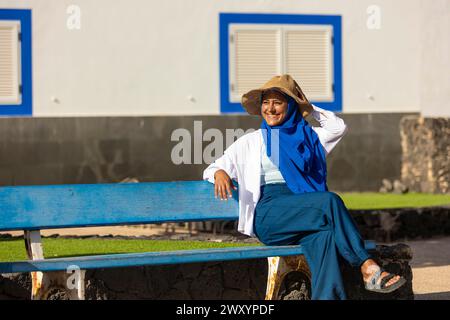 Allegra donna multietnica con un cappello di paglia e sciarpa blu seduta su una panchina blu all'aperto, godendosi una giornata di sole Foto Stock