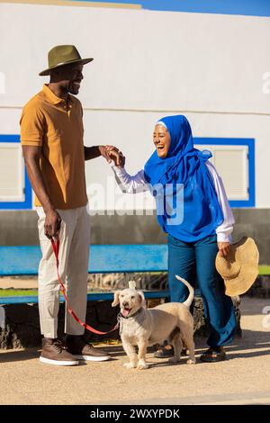 Un momento di gioia quando un uomo con un cappello stringe la mano a una donna in un hijab blu, entrambi sorridendo molto mentre cammina con un cane felice in una giornata di sole Foto Stock