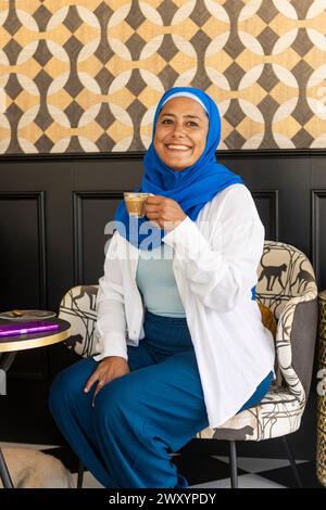 Allegra donna multietnica che indossa un hijab blu sorseggia un caffè in un caffè elegantemente decorato con carta da parati geometrica Foto Stock