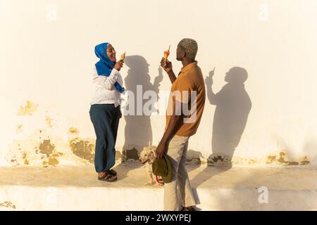 Una coppia multietnica condivide un momento di luce sul gelato contro una parete illuminata dal sole, generando ombre giocose Foto Stock