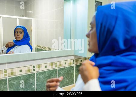 Una donna musulmana premurosa in un hijab blu si riflette in uno specchio del bagno mentre apporta modifiche, rappresentando l'abito culturale e l'identità Foto Stock