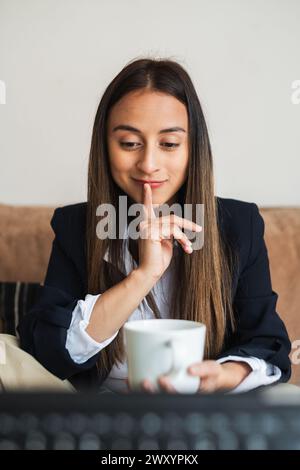 Una giovane donna d'affari siede comodamente, tenendo una tazza da caffè con un'espressione contemplativa, godendosi una pausa al lavoro Foto Stock