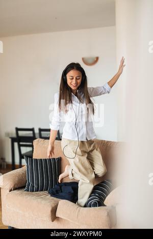Una giovane donna gioiosa con abbigliamento casual e cuffie balla su un divano in un luminoso salotto, esprimendo felicità e libertà Foto Stock