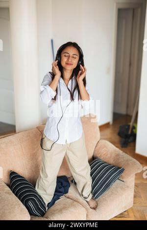 Una giovane donna adulta in piedi su un divano, perduta nella musica con gli occhi chiusi e le cuffie indossate Foto Stock