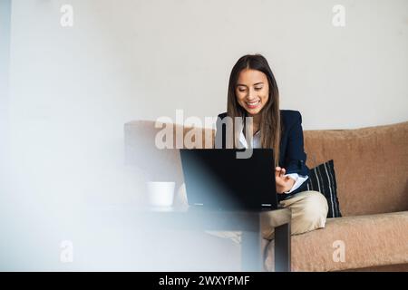 Donna d'affari sorridente che utilizza un computer portatile per una videochiamata mentre si siede su un divano in un ufficio moderno Foto Stock