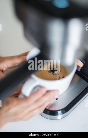 Un'immagine ravvicinata che cattura il momento in cui un espresso fresco viene estratto da una moderna macchina per il caffè in una tazza bianca tenuta da una persona Foto Stock