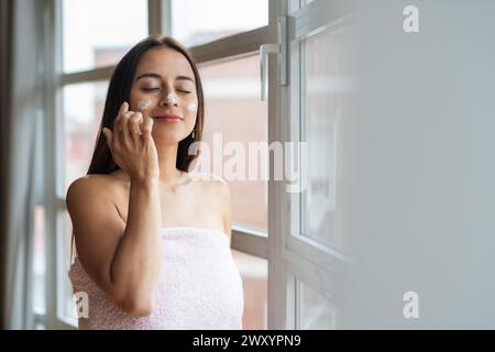 Una donna avvolta in un asciugamano che le applica un idratante sul viso vicino a una finestra luminosa, concentrandosi sulla cura della pelle Foto Stock
