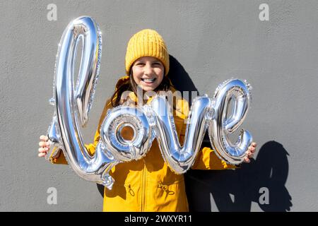 Una donna gioiosa con un cappello giallo e un cappotto sorride mentre regge palloncini in argento lucido a forma di parola amore contro un muro grigio Foto Stock