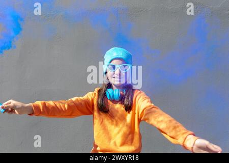 Ragazza sorridente con una felpa arancione vivace, berretto blu e occhiali da sole, con cuffie intorno al collo, in piedi con le braccia allungate Foto Stock