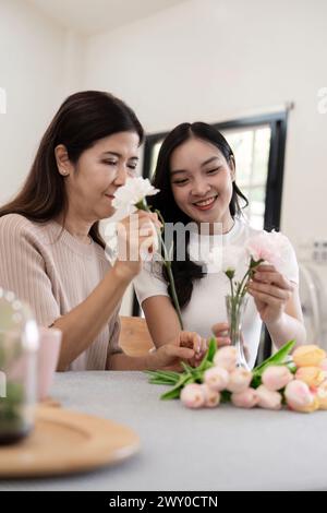 Madre anziana e figlia adulta felici sul tavolo mentre sistemano i fiori in un vaso insieme. Tecnologia e stile di vita. Buon tempo insieme Foto Stock