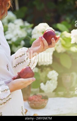 Una donna con una tradizionale blusa ricamata regge delicatamente una mela matura su un tavolo rivestito di pizzi. Vita sostenibile, femminilità e l'eleganza del rurale Foto Stock