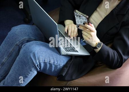 Ragazza in jeans seduta su sgabello imbottito con laptop in ginocchio e smartphone. Lavorare comodamente in ufficio Foto Stock