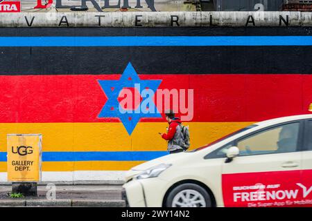 Berlino, Germania. 3 aprile 2024. Una bandiera israeliana si mescola con la bandiera tedesca mentre le persone passano davanti con ombrelli e impermeabili in caso di pioggia: La East Side Gallery di Berlino. Un progetto di graffiti conservato di 118 artisti che copre parte del muro di Berlino e ne celebra la scomparsa. Crediti: Guy Bell/Alamy Live News Foto Stock