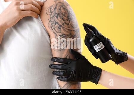 Maestro del tatuaggio che spruzza il braccio dell'uomo su sfondo giallo, primo piano Foto Stock