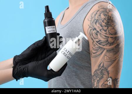 Maestro del tatuaggio con bottiglie di prodotti cosmetici e uomo su sfondo blu, primo piano Foto Stock