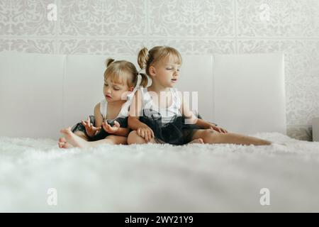Due ragazze sono sedute fianco a fianco su un letto bianco, guardando qualcosa fuori dalla telecamera. Indossano un abbigliamento casual e sembrano avvolti da wha Foto Stock