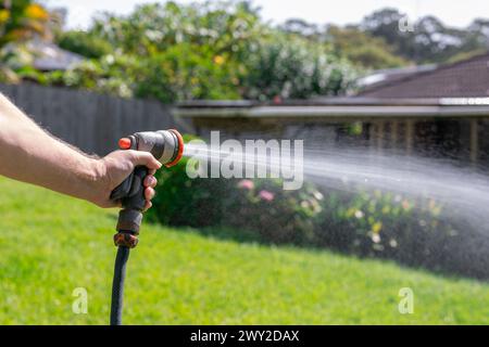 Tubo da giardino con ugello regolabile. Mano dell'uomo che tiene la pistola a spruzzo e innaffia le piante, spruzzando acqua sull'erba nel cortile. Foto Stock