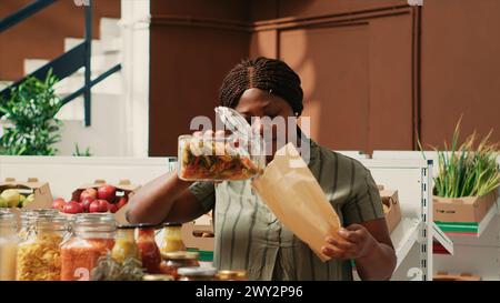 Cliente donna che riempie il sacchetto di carta con pasta venduta come articolo sfuso, acquistando vari tipi di prodotti senza additivi biologici dal mercato agricolo locale. Acquirente in cerca di forniture per dispensa biologica. Foto Stock