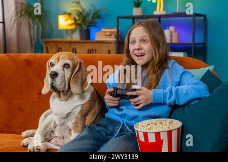 Eccitata giovane ragazza felice che usa il controller joystick che gioca con la console video gioco televisivo divertendosi a sedersi sul divano in camera. Ragazzo preadolescente caucasico che si diverte a giocare online a casa con il cane beagle Foto Stock