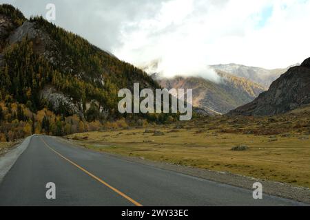 Una strada asfaltata a due corsie si trasforma in un'ampia fessura verso alte montagne con cime tra le nuvole in una soleggiata giornata autunnale. Chuisky Tract, Altai, Siberia Foto Stock