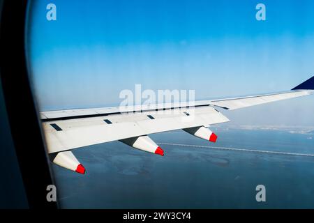 Ala di aerei con la punta rossa in volo sopra la costa oceanica con edifici cittadini e ponte appena visibile sotto.g che volano sopra il ponte Foto Stock