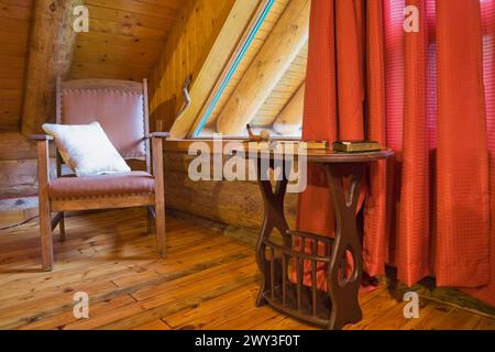 Poltrona in legno marrone con cuscino bianco e tavolino nella camera da letto principale con tende rosse e pavimento in legno al piano superiore all'interno Foto Stock