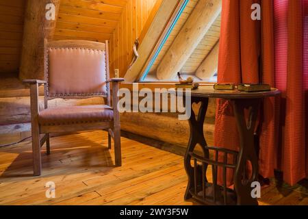 Poltrona in pelle di legno marrone e tavolino nella camera da letto principale con tende rosse e pavimento in legno al piano superiore all'interno rustico contemporaneo Foto Stock