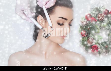 Il cosmetologo fa la procedura di terapia Microcurrent di una bella donna in un salone di bellezza. Faccia da donna ravvicinata e corona di Capodanno con la neve Foto Stock