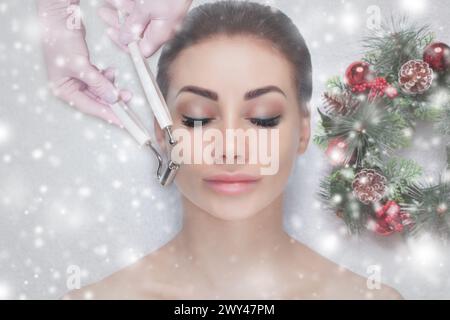 Il cosmetologo fa la procedura di terapia Microcurrent di una bella donna in un salone di bellezza. Faccia da donna ravvicinata e corona di Capodanno con la neve Foto Stock