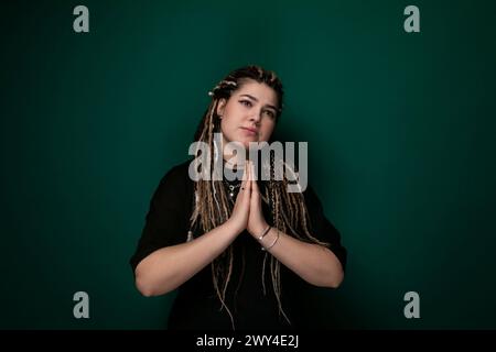 Una donna con i dreadlock si trova in posizione verticale davanti a una parete verde fissa, guardando direttamente la fotocamera. Ha un'espressione calma sul viso e.. Foto Stock