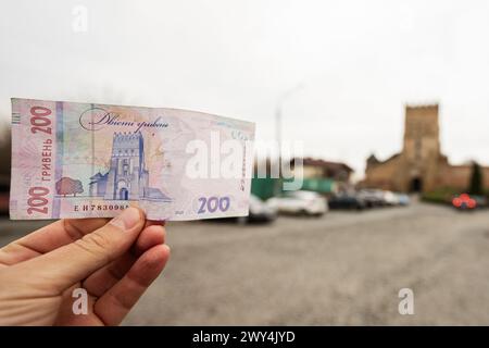 Lutsk, Ucraina - marzo 2024: Primo piano di una mano che espone una banconota Ucraina del 200 con un castello storico sfocato e un cielo nuvoloso nella parte posteriore Foto Stock