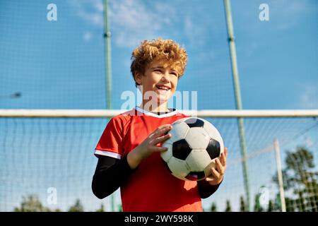 Un ragazzino si trova di fronte a una porta di calcio, tenendo un pallone da calcio con un'espressione determinata. E' posizionato per un calcio, mostrando il suo amore per Foto Stock