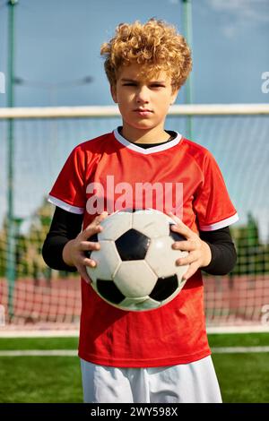 Un ragazzino si trova in un vasto campo da calcio, cullando un pallone da calcio vicino al petto. L'erba verde brillante si estende all'infinito intorno a Hi Foto Stock