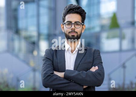 Ritratto di un giovane uomo d'affari indiano sorridente e di successo in una tuta in piedi fuori per strada, che si incrocia le braccia sul petto e guarda con sicurezza la fotocamera. Foto Stock