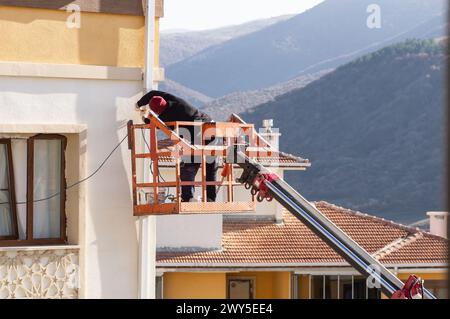 Lavoratore senza casco che ripara il tubo dell'acqua sulla gru, avvitare con un trapano Foto Stock