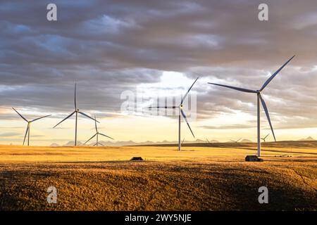 Fila di mulini a vento che generano elettricità sulle praterie canadesi che si affacciano su un paio di vecchi capannoni di fattoria sotto un suggestivo cielo al tramonto vicino a Pincher Creek A. Foto Stock