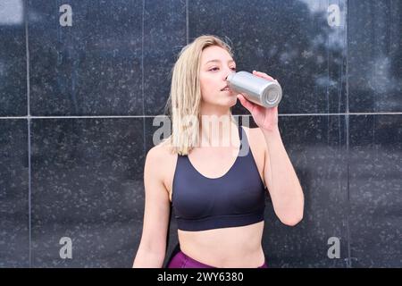 giovane donna bionda con una maglia sportiva che beve acqua su sfondo neutro Foto Stock