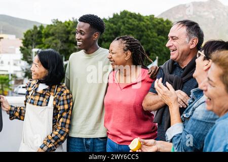 Amici felici e multigenerazionali che si divertono a preparare il cibo sul tetto della casa - diversità persone stile di vita Foto Stock