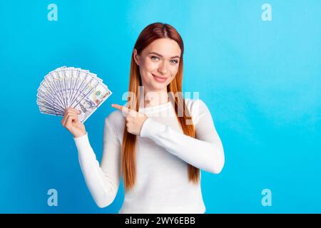 Ritratto fotografico di una giovane donna attraente che tiene i soldi del denaro a punta vestiti eleganti bianchi isolati su sfondo di colore blu Foto Stock