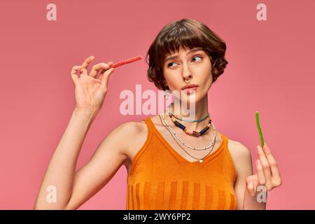 dubbiosa ragazza trafitto sui vent'anni mangiando due gusti diversi di caramelle dolci e acide sul rosa Foto Stock