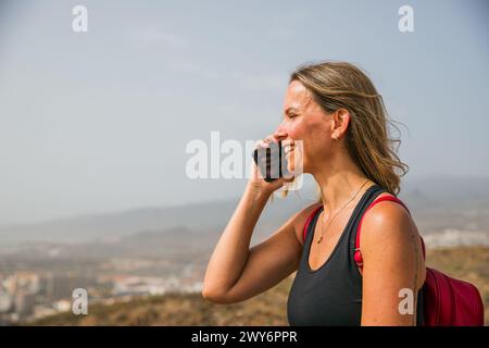 Una donna sta parlando al cellulare mentre si trova su una collina. Sta sorridendo e si sta godendo la conversazione Foto Stock