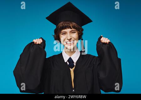 giovane bruna laureata in berretto accademico che si sente felice su sfondo blu, successo Foto Stock