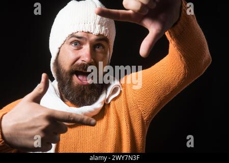 L'uomo felice con maglione arancione, cappello bianco e sciarpa rende la cornice della fotocamera con le dita. Uomo con barba che utilizza l'inquadratura manuale per creare il mirino. Immaginario Foto Stock